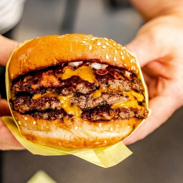 Hands holding a double cheeseburger with melted cheese, bacon, and onions on a sesame seed bun.