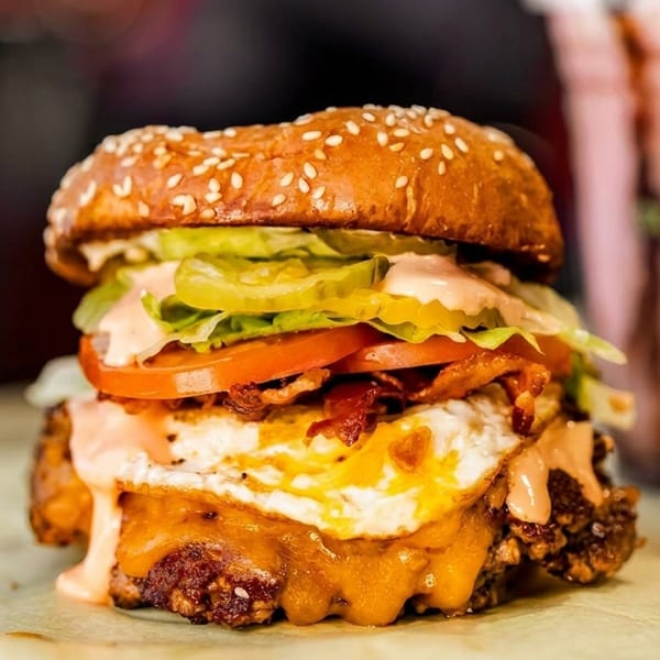 A loaded burger with fried egg, bacon, cheese, lettuce, tomato, and sauce, with a milkshake in the background.